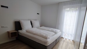 A modern bedroom with a large bed, a bedside table, and a window with sheer curtains. - Smart & Stay Aparthotel (Saarlouis)