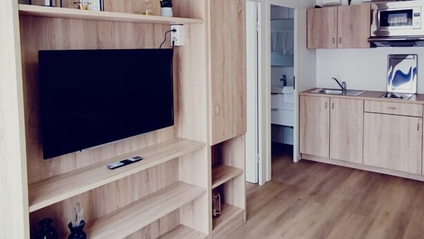 A modern living room with a flat-screen TV, wooden shelving, and a kitchen area with cabinets and appliances. - Smart & Stay Aparthotel (Saarlouis)