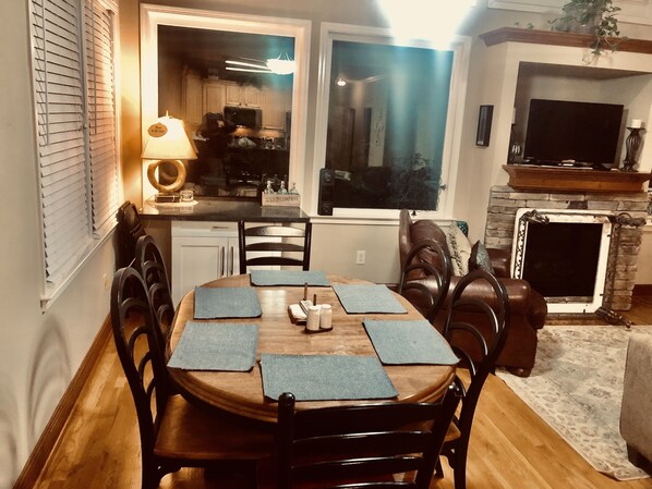 A dining area with a wooden table, black chairs, and blue placemats. There is a kitchen visible through an open door, a fireplace, and a television mounted above it. - Chalet Suzanne of Helen (Helen)