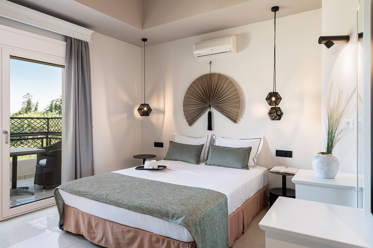 Ein Schlafzimmer mit Bett, Nachttischen, einem Ventilator, einem Fenster mit Vorhängen und Blick ins Freie.
