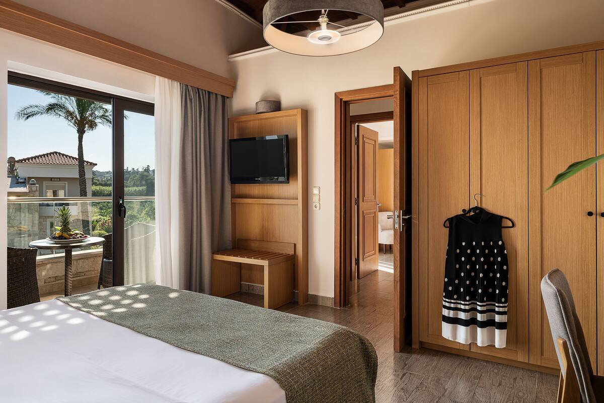 Ein Hotelzimmer mit Bett, einem an einer Holzeinrichtung montierten Fernseher, einem Balkon mit Tisch und Stühlen und Blick auf Palmen sowie ein Haus.