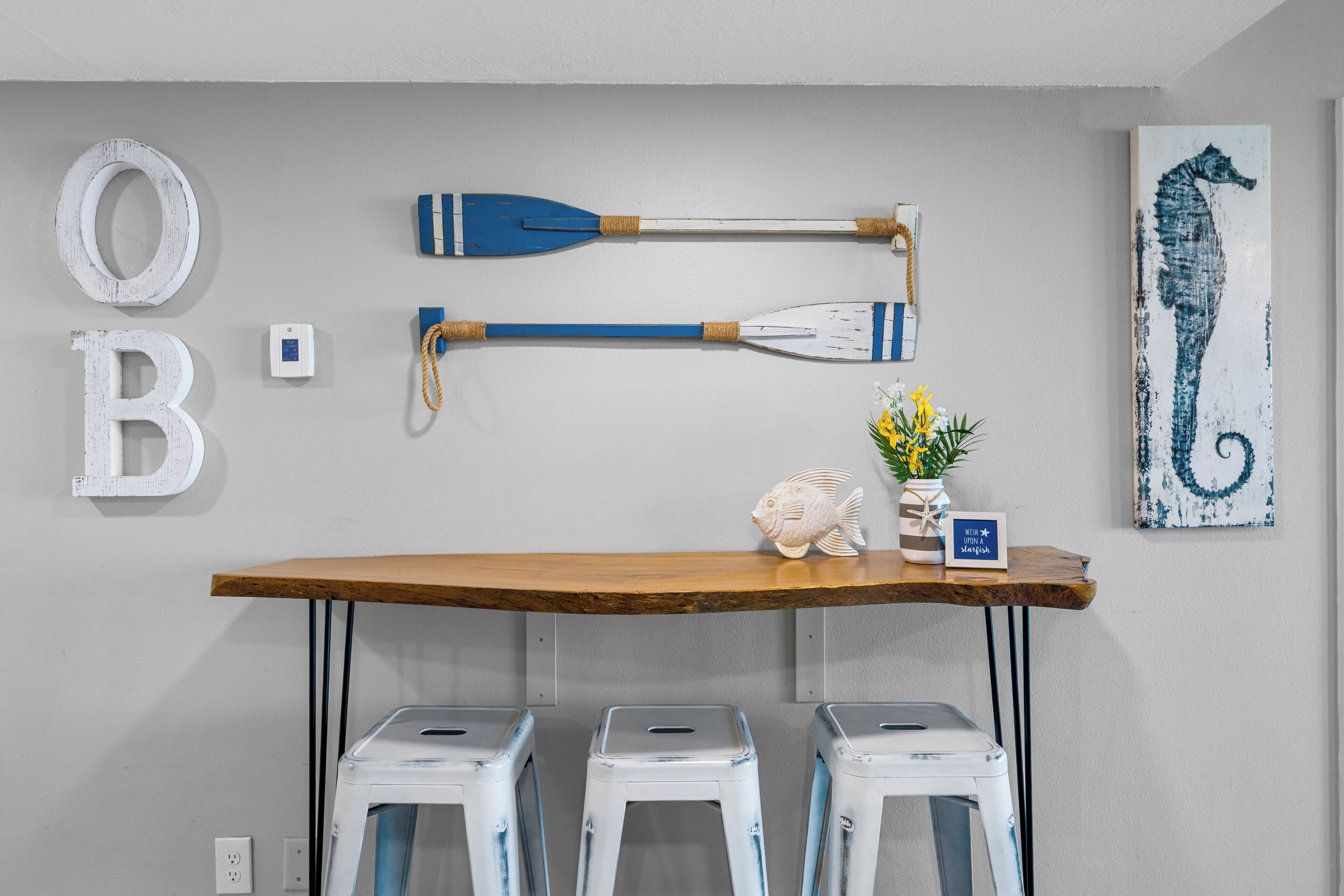 A room with a wooden table, three white stools, a wall-mounted paddle, and a seahorse painting.