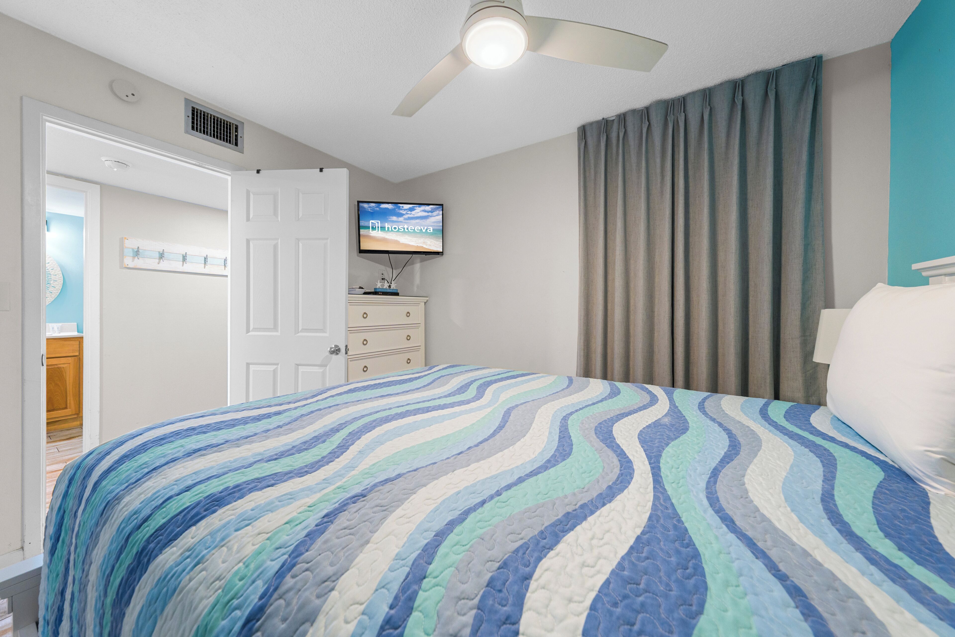 A bedroom with a bed, a ceiling fan, a television, and a door leading to another room.