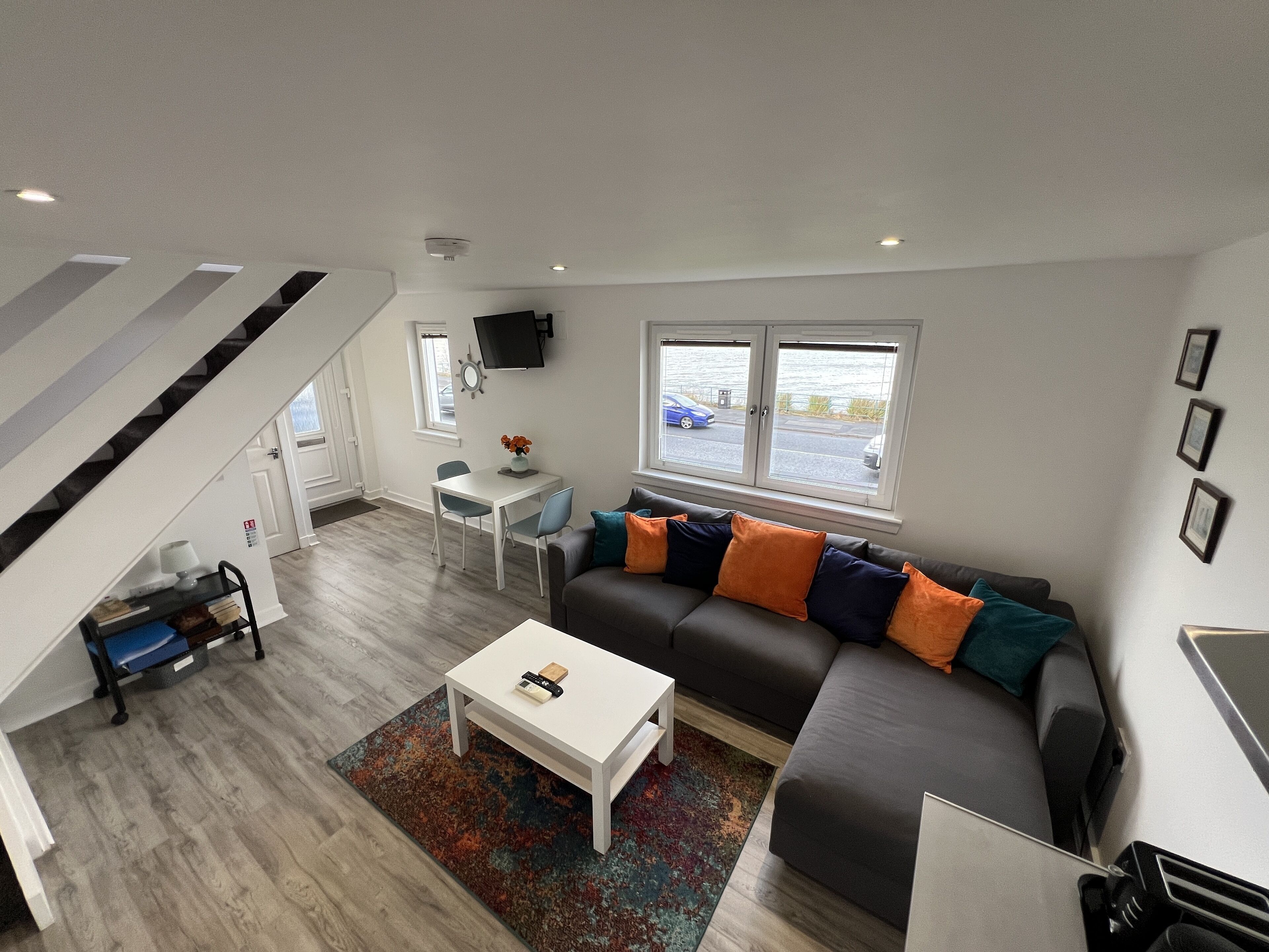 Un salon moderne avec un canapé d’angle gris, une table basse blanche et un tapis coloré. On y trouve également un coin repas avec une table et des chaises blanches, et une cuisine en arrière-plan.