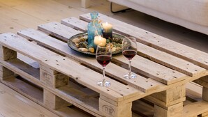 A cozy living room with a wooden pallet coffee table, a white sofa with a teal pillow, a small side table with a lamp, and a vase with dried plants. - A private Villa infront of the sea (Paros Island)