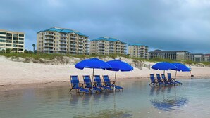 A beach with blue umbrellas and lounge chairs, a sandy shore, and buildings in the background. - Luxury 5th Floor Oceanfront Condo next to Ritz Carlton! (Fernandina Beach)