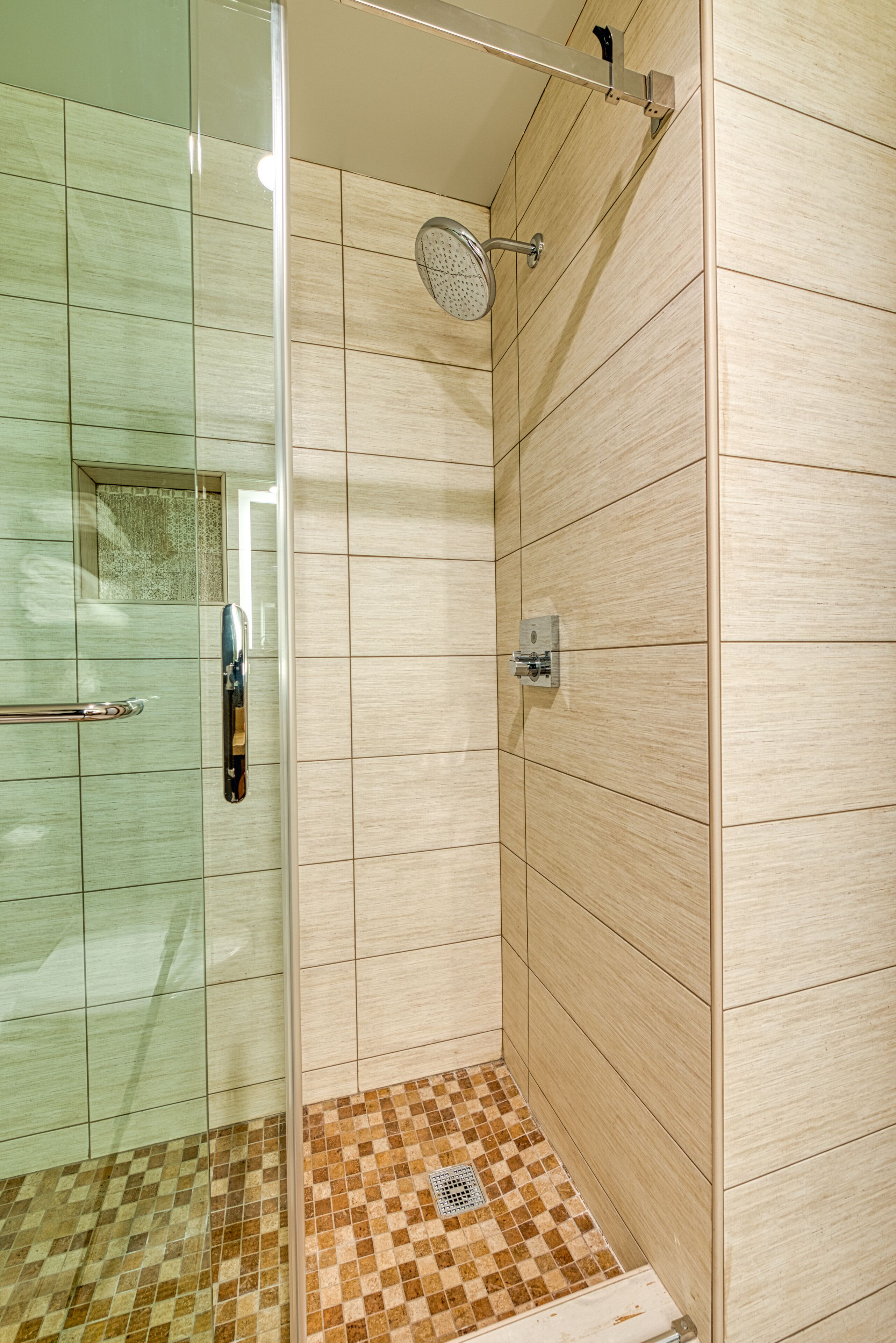 Una ducha con mampara de vidrio, un cabezal de ducha manual, paredes de azulejos y piso de diseño de cuadros.