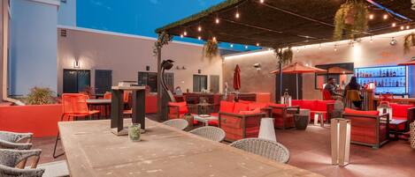 An outdoor seating area with wicker furniture and red cushions, a bar with bottles, and a covered section with string lights.