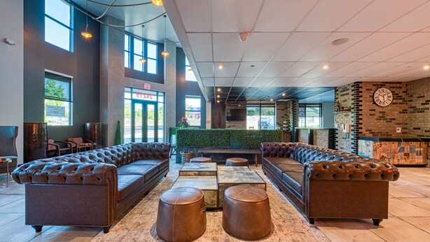 A spacious lobby with leather sofas, ottomans, and a reception area with a green wall and brick accents. - Hotel Lexen Santa Clarita Valencia Near Six Flags (Newhall)
