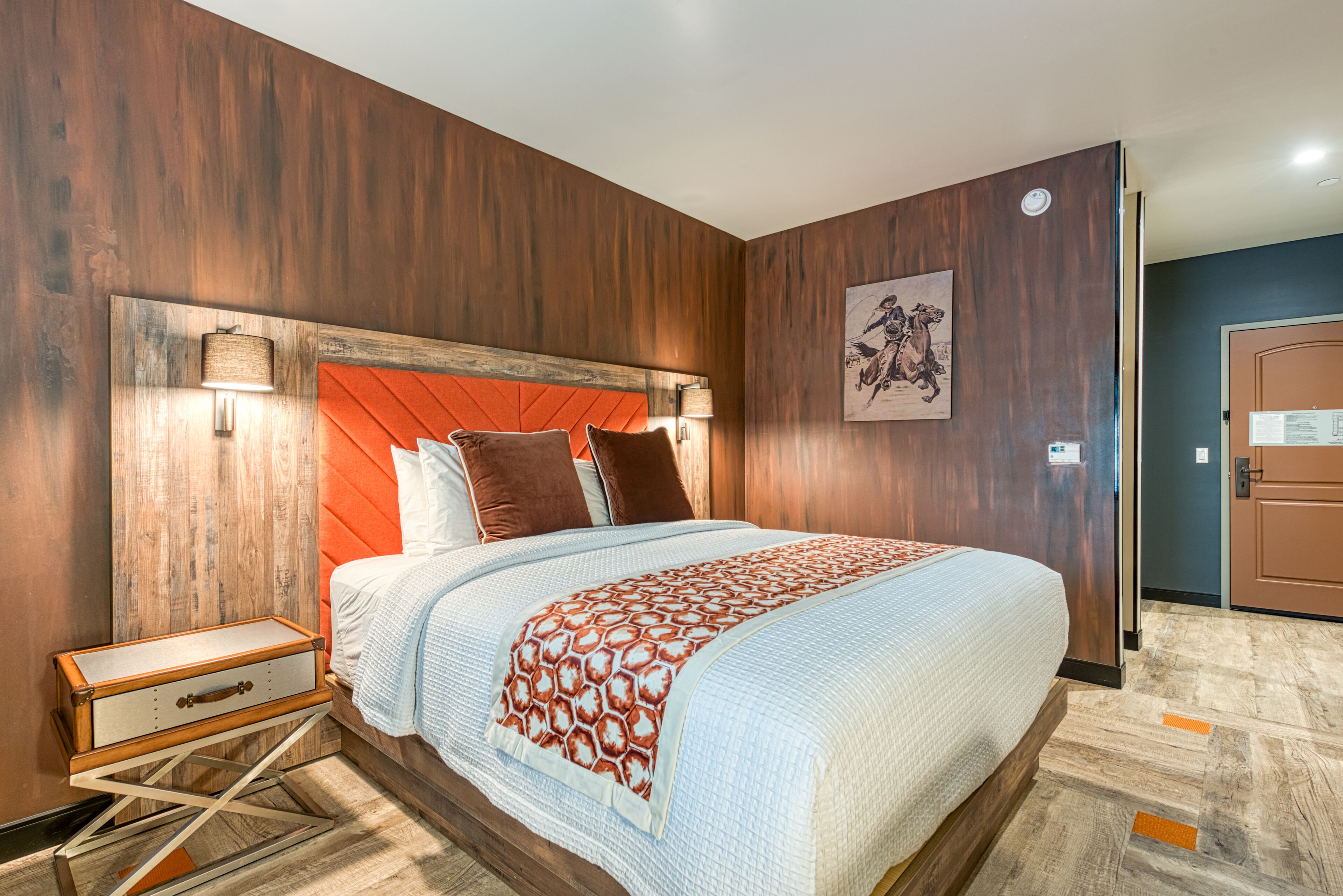 Ein Hotelzimmer mit einem hölzernen Kopfteil, einem Bett mit gemustertem Überwurf, einem Nachttisch und einem Wandbild.