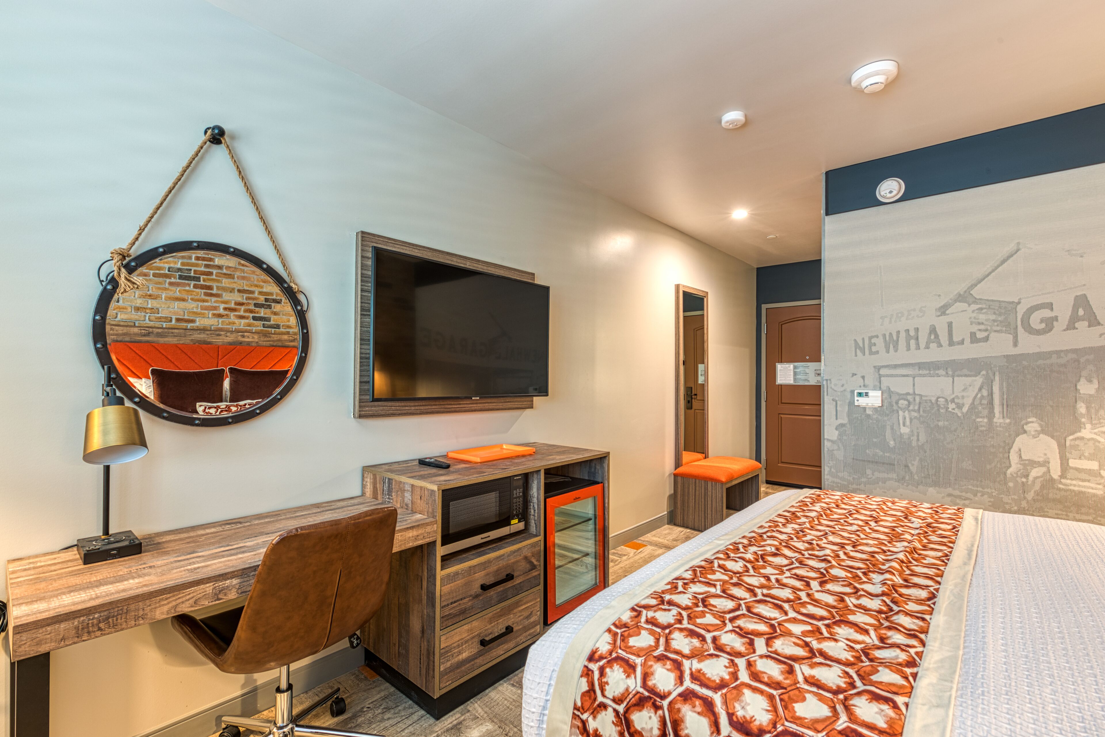 Habitación de hotel moderna con un escritorio de madera, una silla cómoda, una cama con colcha estampada, un televisor de pantalla plana y un espejo fijado a la pared.