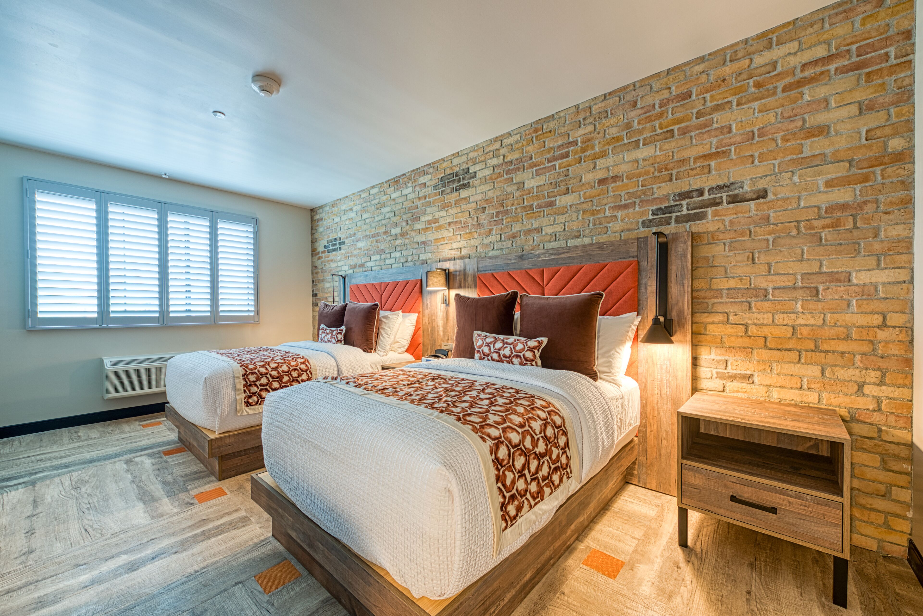 Habitación doble con pared de ladrillo, dos camas, una mesita de noche y una ventana con persianas.