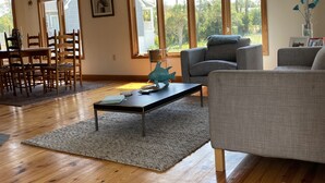 Un salón amplio con suelo de madera, un sofá gris, una mesa de comedor con sillas y grandes ventanales que ofrecen vistas al verdor.