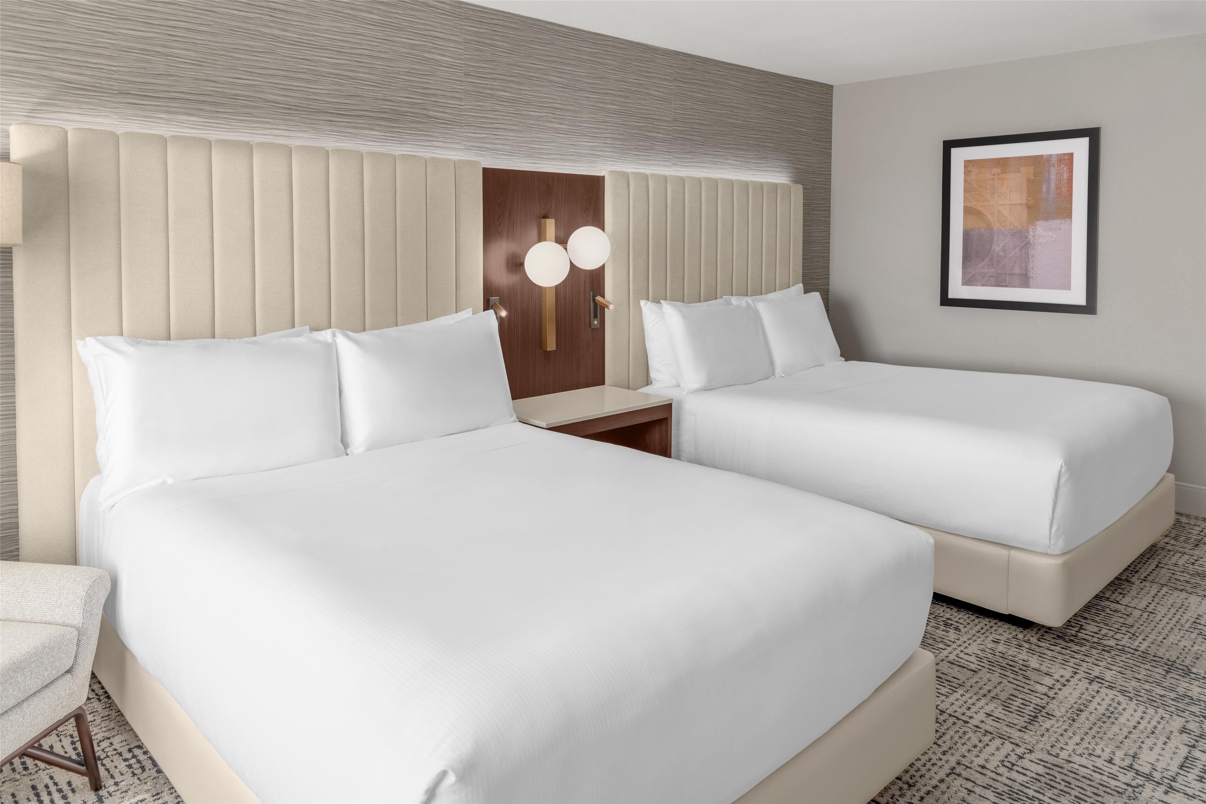 A hotel room with two beds, a wooden door, a framed picture on the wall, and a small table with a lamp.