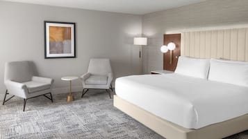 Una habitación de hotel con una cama grande, dos sillones, una mesita redonda, una lámpara de pared y una obra de arte enmarcada.