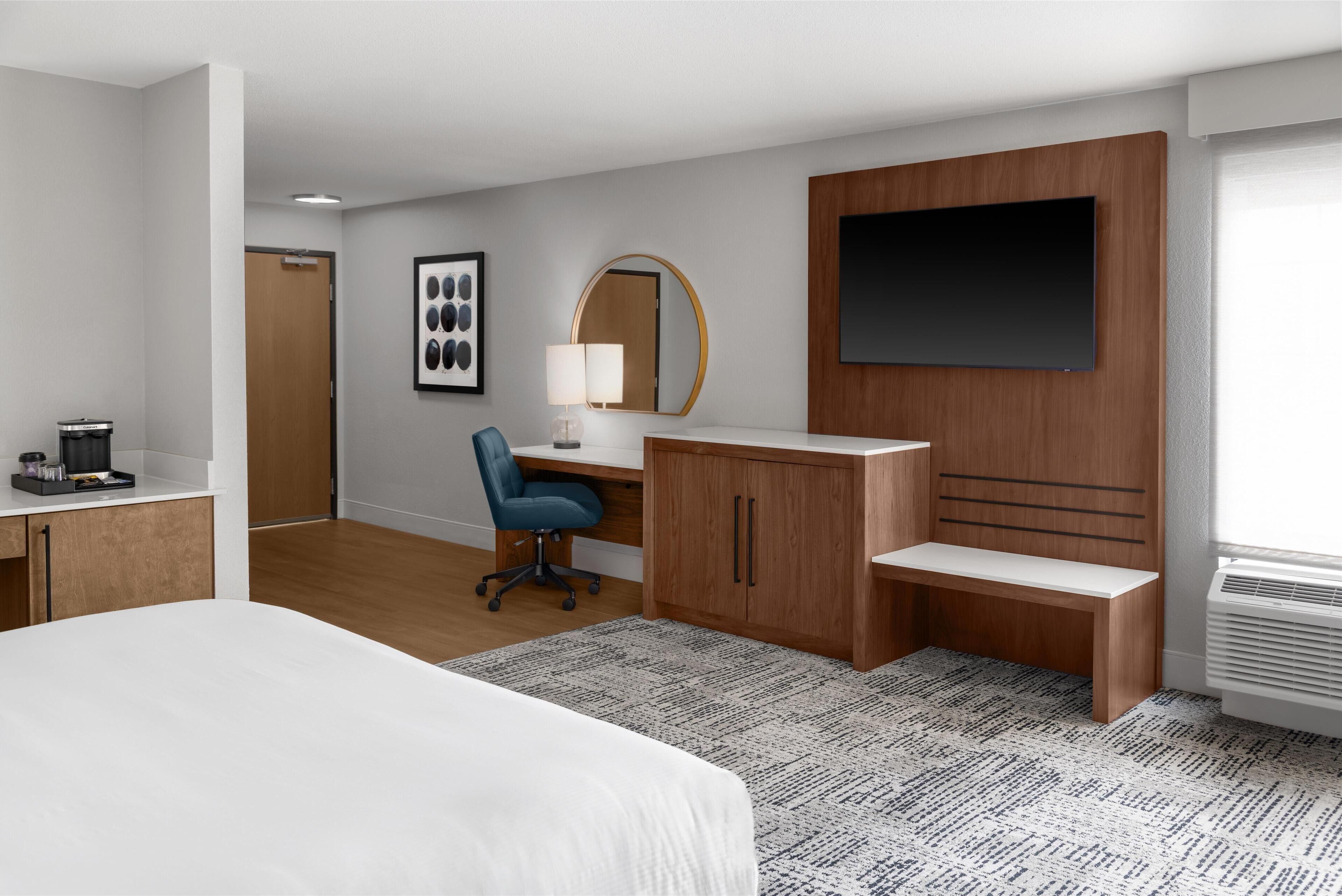 A hotel room with a bed, desk, chair, TV, and a large mirror.