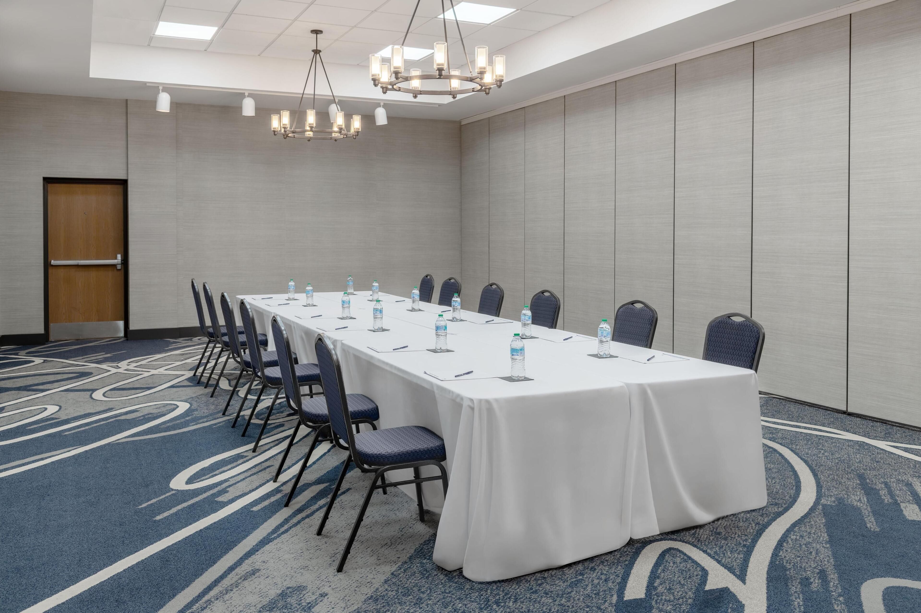 Una sala conferenze con un lungo tavolo, sedie, bottiglie d'acqua e un lampadario.