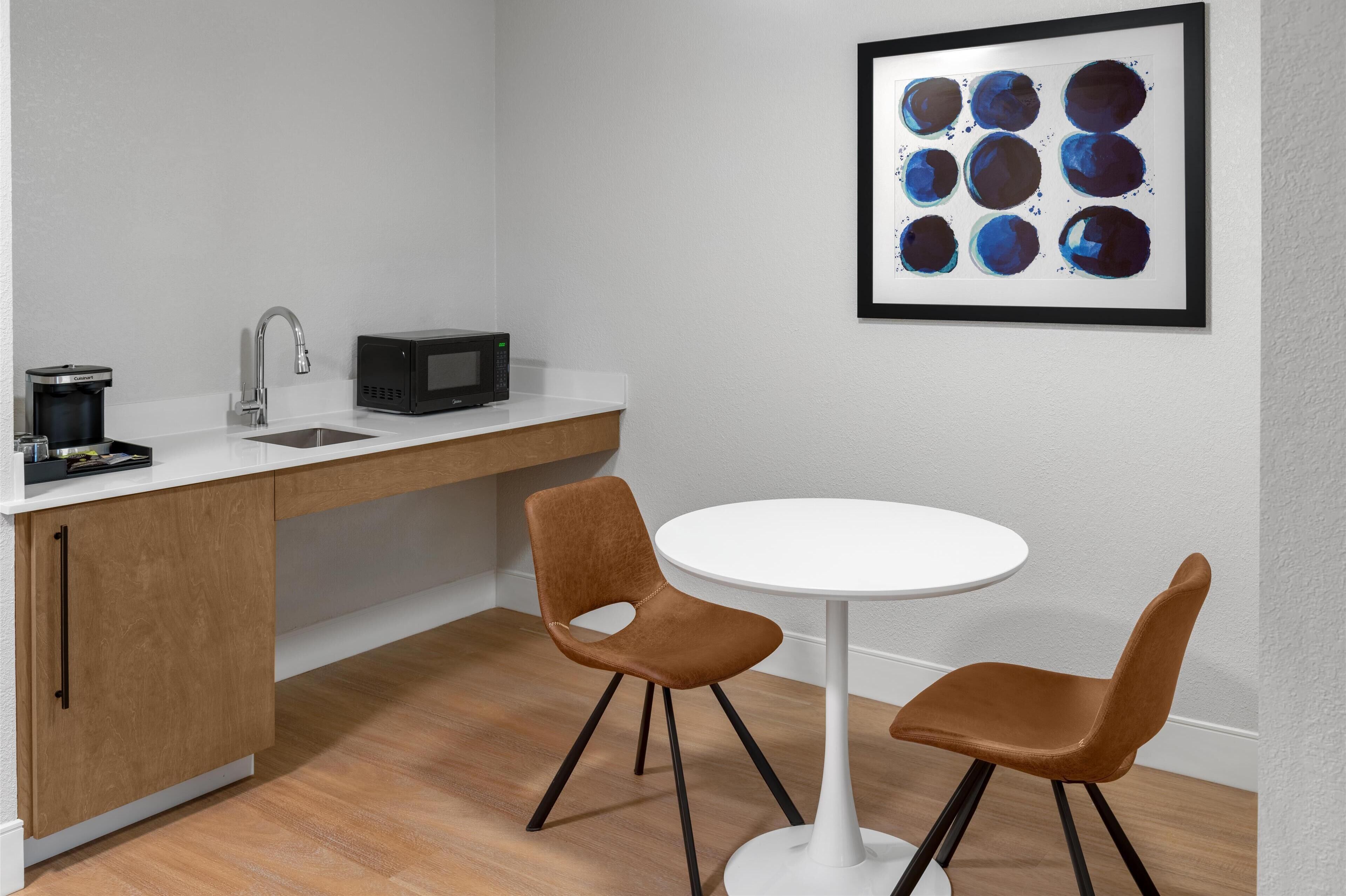 A modern kitchen with a white countertop, a sink, a microwave, and a coffee maker. There is a round white table with two brown chairs. A framed abstract painting is on the wall.