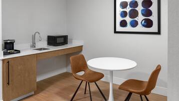 A modern kitchen with a white countertop, a sink, a microwave, and a coffee maker. There is a round white table with two brown chairs. A framed abstract painting is on the wall.