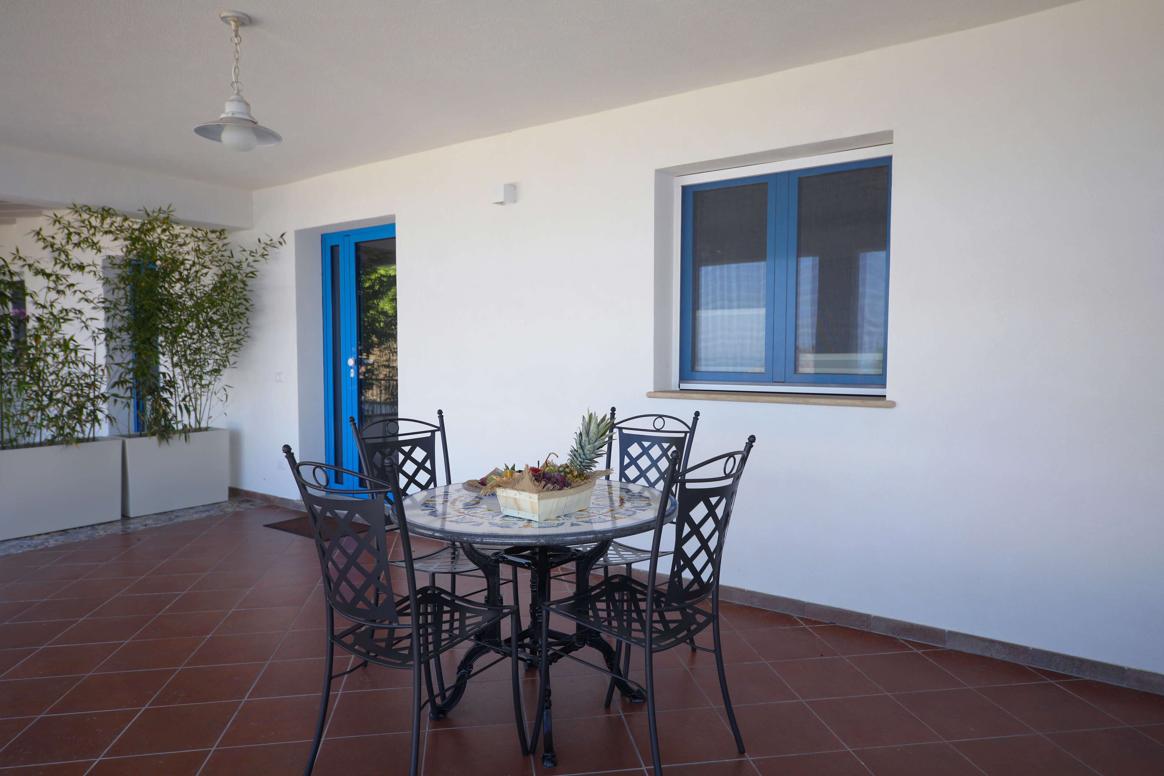A dining area with a round table and four chairs, a bowl of fruit, and a window with blue trim.