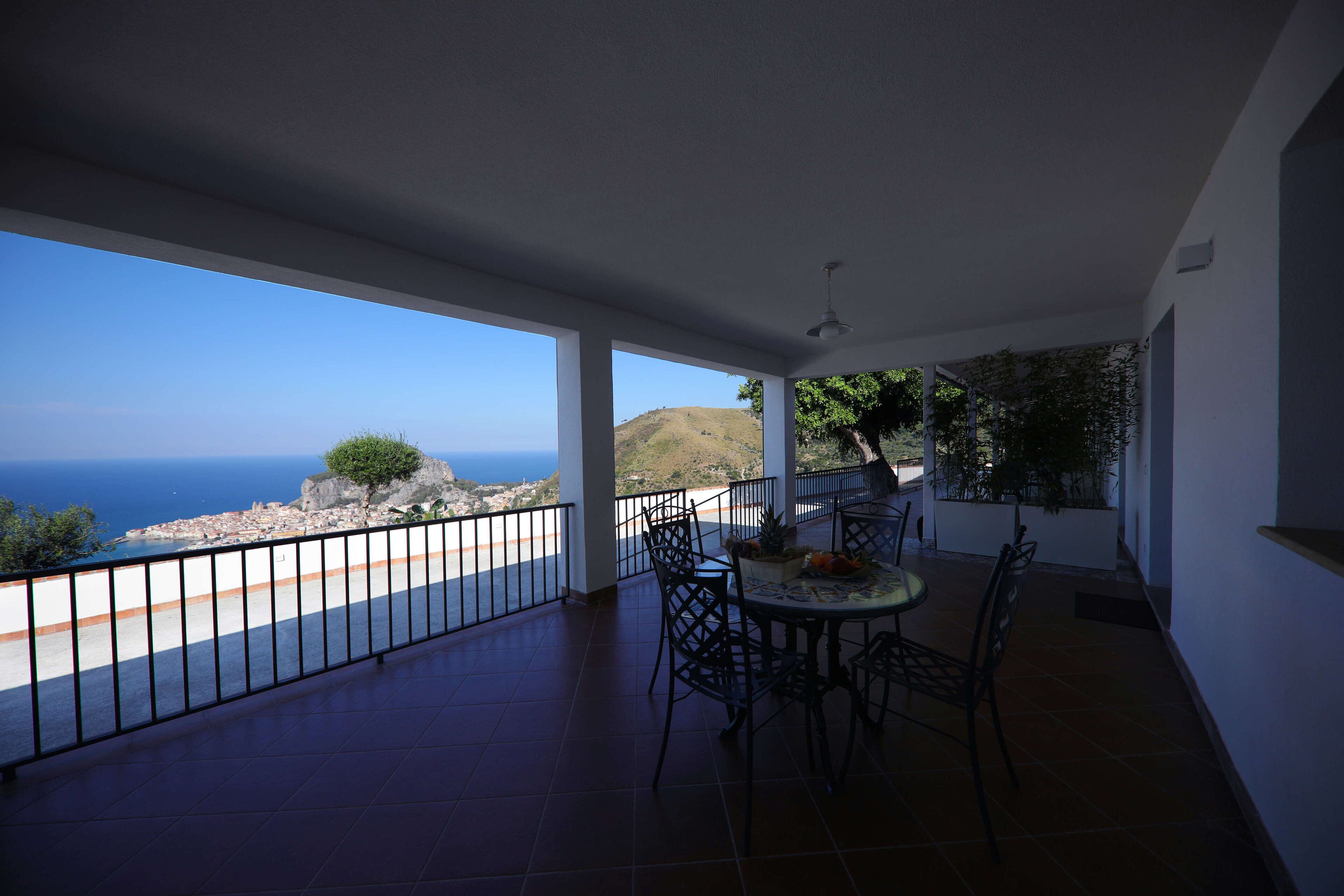 A balcony with a table and chairs, overlooking a coastal view with a distant hill and sea.