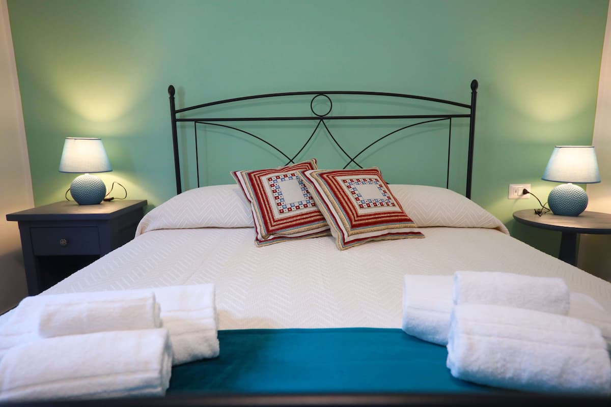 A neatly made bed with white linens and two decorative pillows, flanked by two bedside tables with lamps, in a room with green walls.