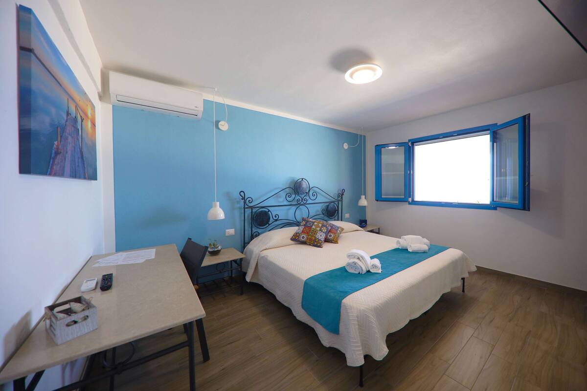 A bedroom with a bed, a desk, a chair, a window, and a painting on the wall.