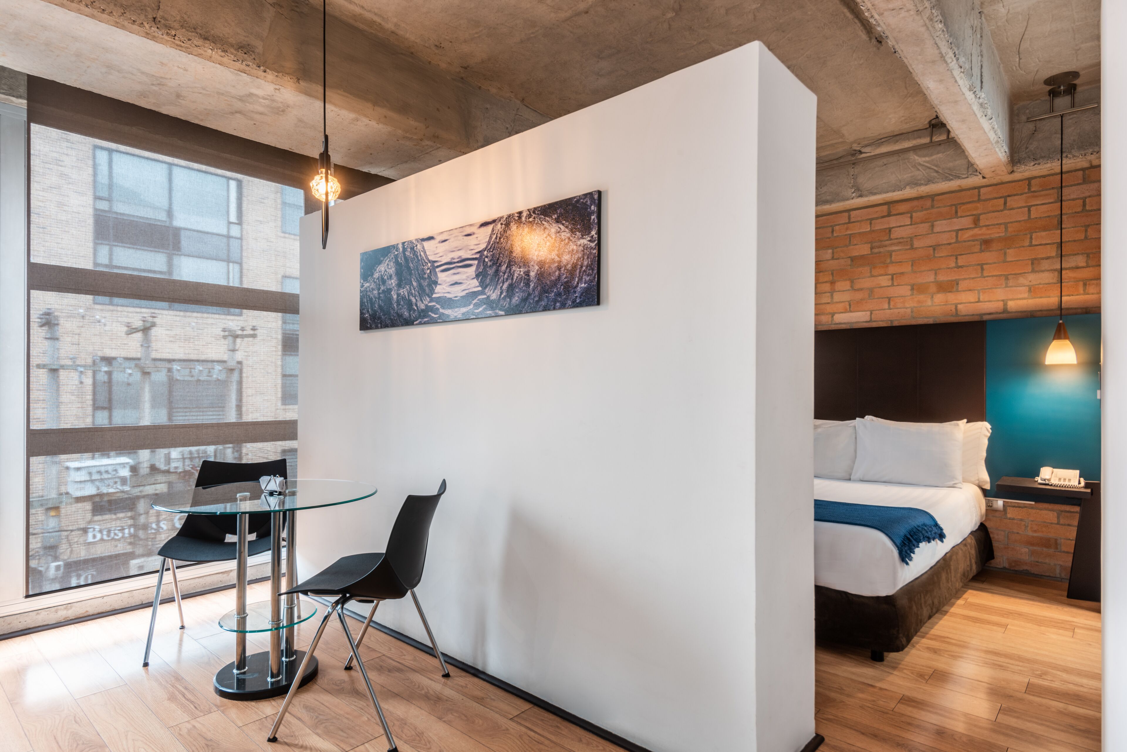 A modern hotel room with a large window, a bed with a blue blanket, a desk with two chairs, and a painting on the wall.