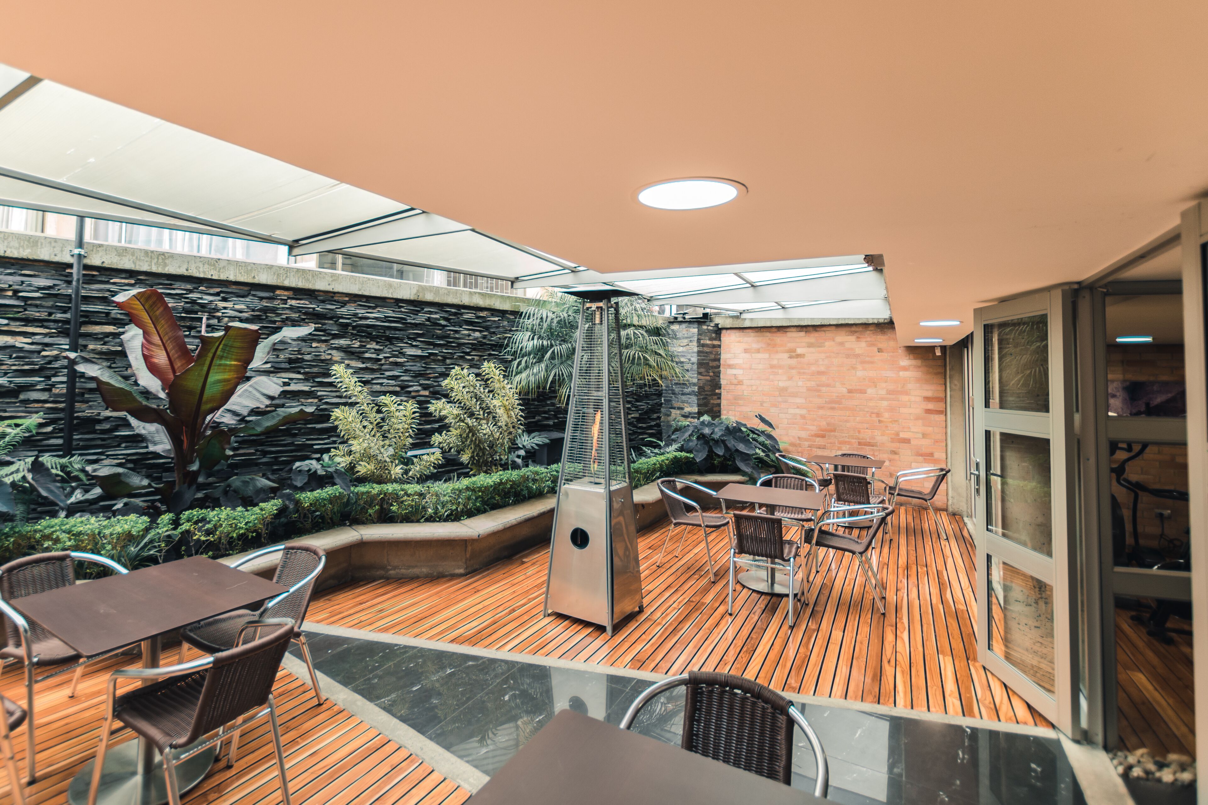 An outdoor patio area with wooden flooring, metal chairs, a glass table, and a stone wall with plants.