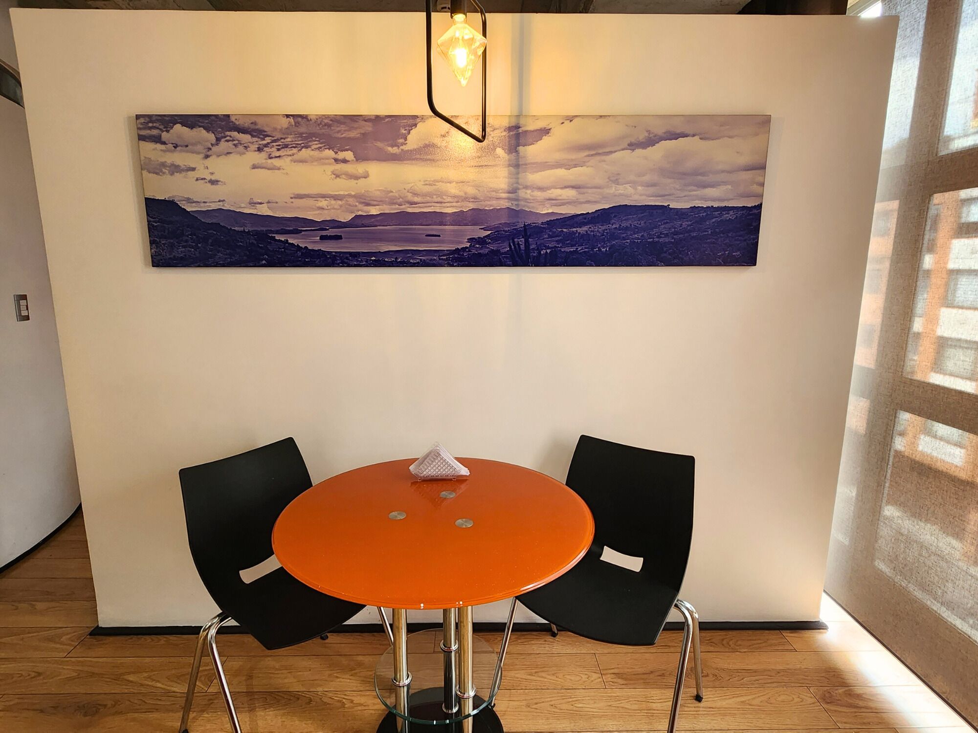 A room with a round orange table and two black chairs, a painting of a landscape on the wall, and a hanging light fixture.