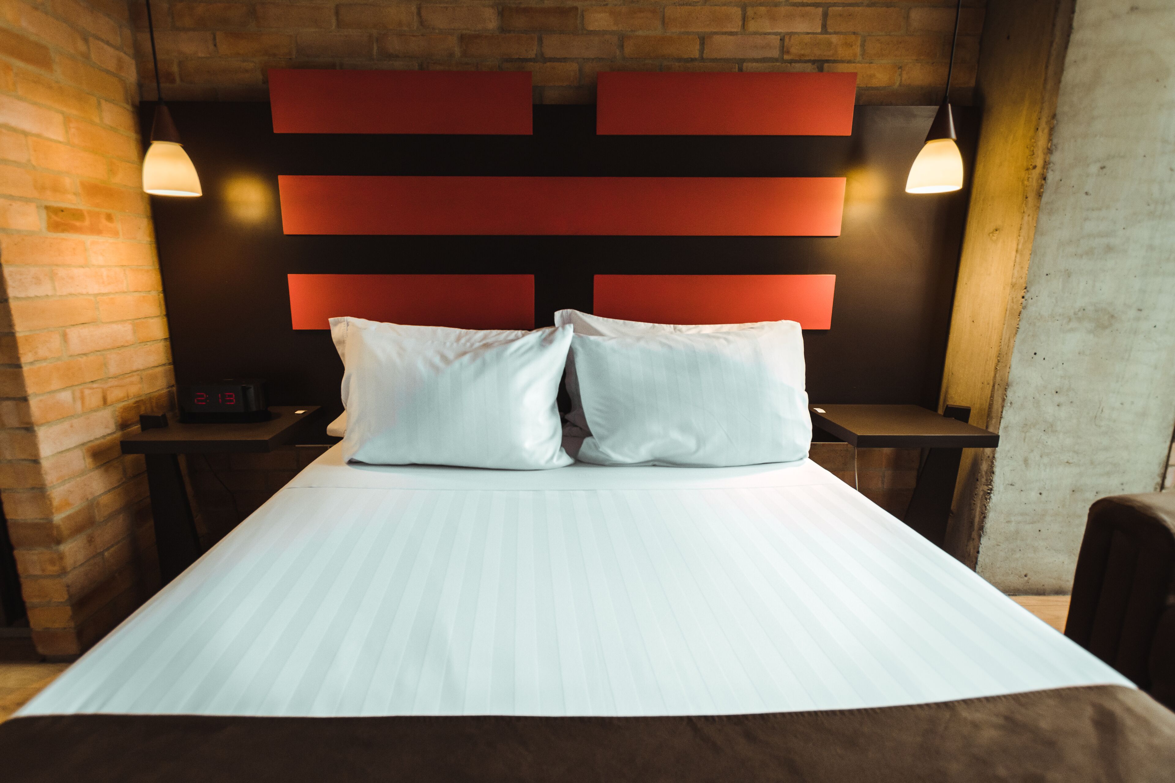 A hotel room with a bed, a brick wall, a red headboard, and two pendant lights.