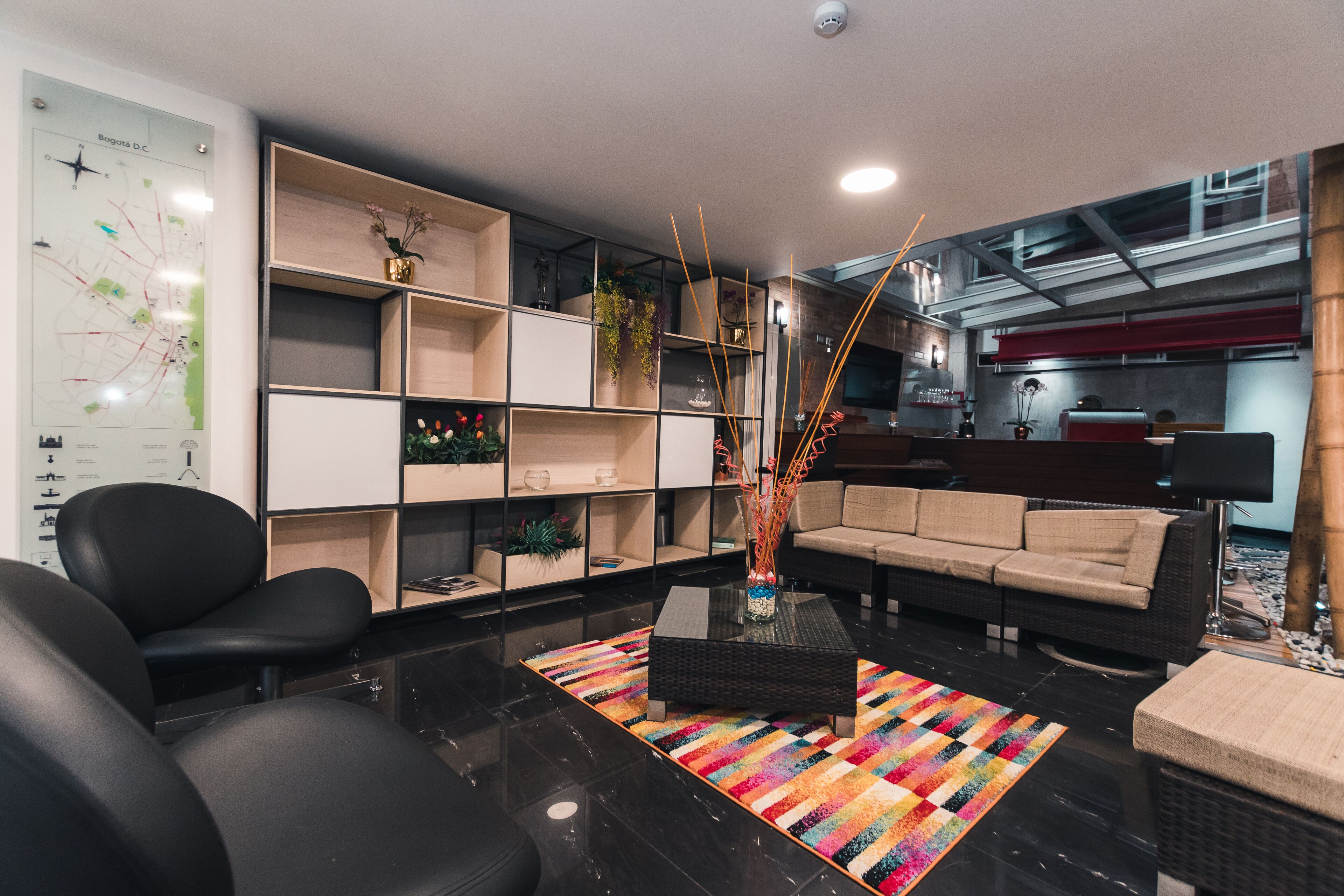 Una sala de estar moderna con un sofá de cuero negro, un sofá beige, una mesa de centro y una alfombra colorida. Hay un mapa en la pared y una estantería con varios objetos.