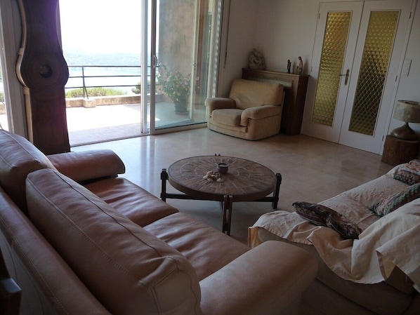 A living room with a sofa, armchair, and round coffee table. There is a sliding glass door leading to a balcony with a view.