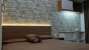 Un dormitorio con cabecera de piedra, cama con cubrecama color beige, escritorio con silla y ventana con cortinas.