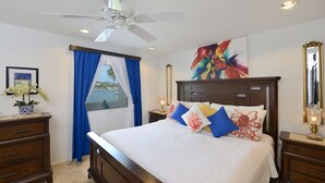 A bedroom with a bed, a ceiling fan, a nightstand with a lamp, a dresser, a window with curtains, and a painting on the wall. - Condo SeaStar 24 (Oyster Pond)
