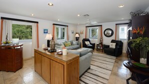 A spacious living room with a wooden coffee table, a grey sofa, and a black leather armchair. There are multiple windows with curtains, a ceiling fan, and a view of greenery outside. - Condo SeaStar 24 (Oyster Pond)