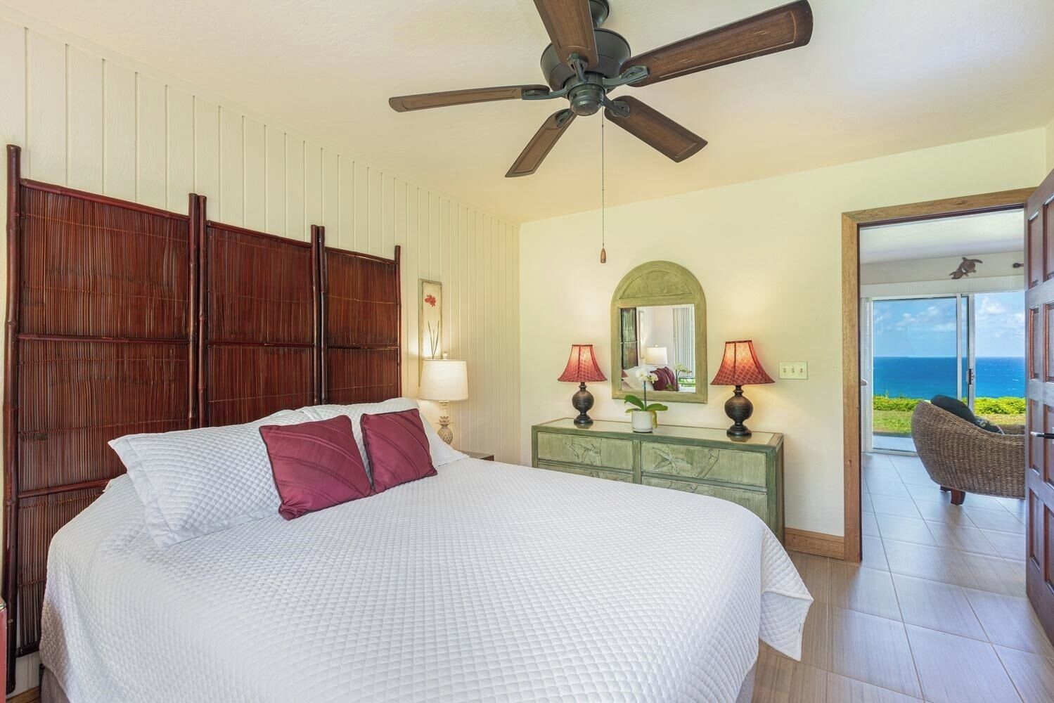A bedroom with a bed, a ceiling fan, a dresser, two lamps, a mirror, and a view of the ocean.