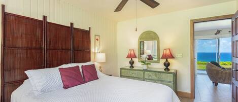 A bedroom with a bed, a ceiling fan, a dresser, two lamps, a mirror, and a view of the ocean.