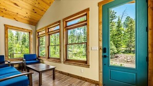 Ein Zimmer mit Holzboden, einer türkisblauen Tür und großen Fenstern mit Blick auf einen Wald.