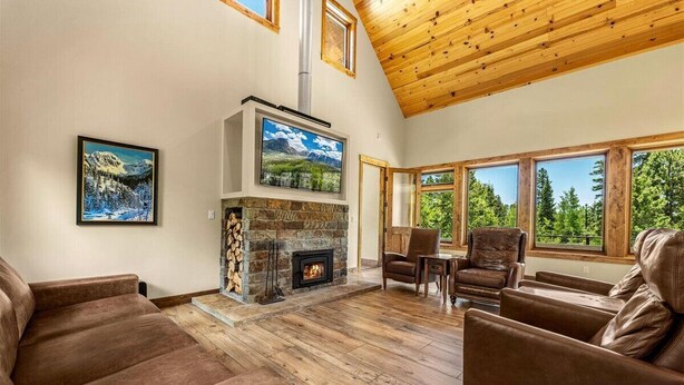 Ein gemütliches Wohnzimmer mit Kamin aus Stein, Ledersofas und großen Fenstern mit Blick auf Bäume.