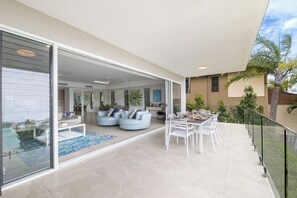A modern living space with a glass sliding door leading to a balcony, a dining area with white furniture, and a pool visible through the glass.