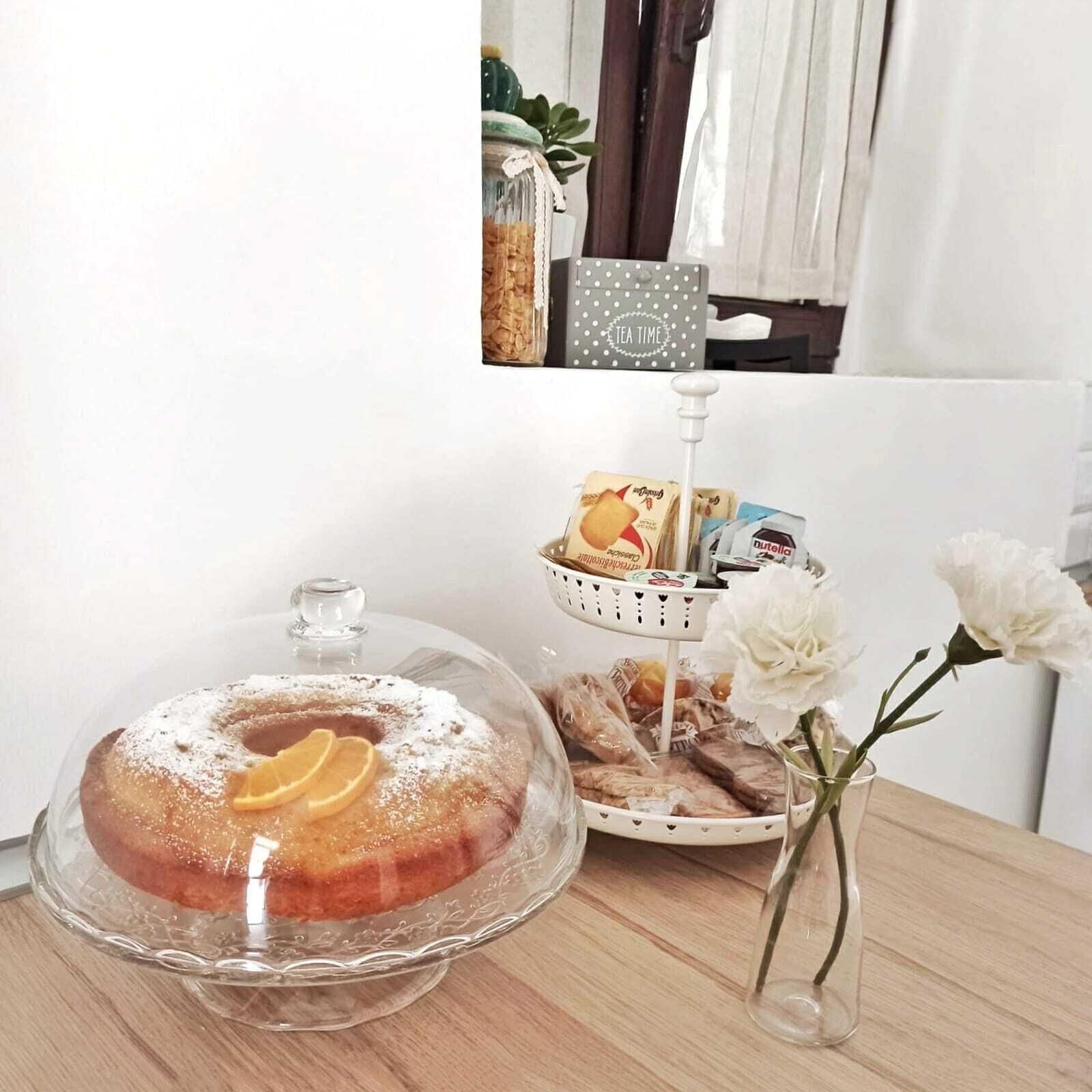 A dessert display with a covered cake, a basket of bread, and a vase with flowers on a wooden table.