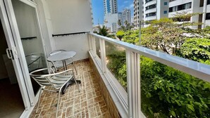 Un balcon doté d’un sol carrelé, d’une petite table et de deux chaises. Il offre une vue sur un jardin où poussent différentes espèces de plantes, ainsi que sur un paysage urbain avec de hauts immeubles en arrière-plan.