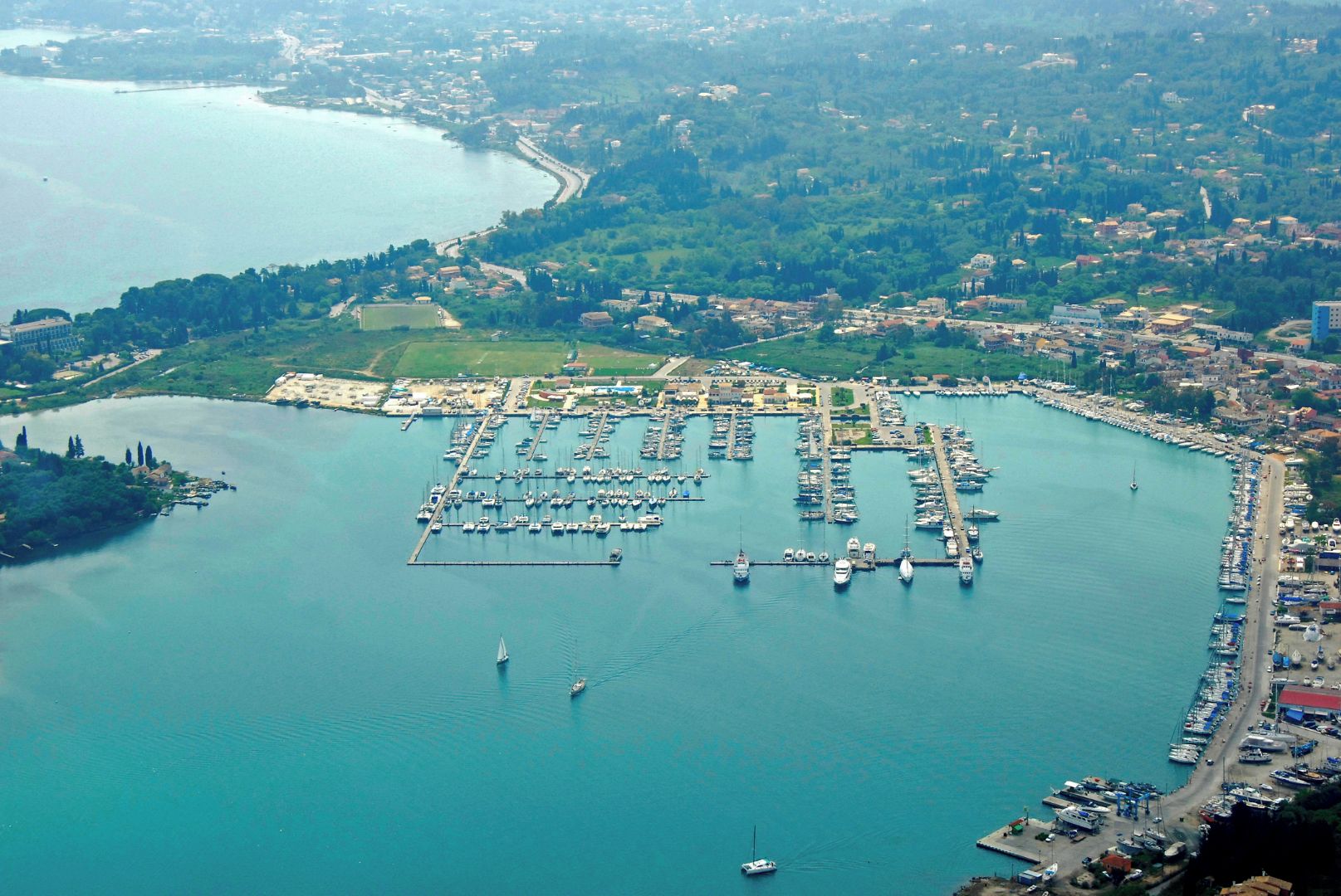 An aerial view of a marina with numerous boats docked, surrounded by a coastal town and greenery.