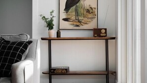 Una sala de estar con un sofá gris, una obra de arte enmarcada de un pájaro, una mesita con una planta y una estantería.