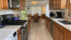 A kitchen with wooden cabinets, a stove, a dishwasher, and a dining area with a table and chairs. - JD36: Flyway | Multiple Decks with Amazing Marsh Views (Ocracoke)