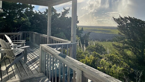 A wooden deck with chairs, overlooking a forested landscape with a clear sky and sun shining through clouds. - JD36: Flyway | Multiple Decks with Amazing Marsh Views (Ocracoke)