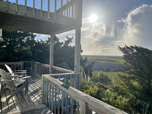 A wooden deck with chairs, overlooking a forested landscape with a clear sky and sun shining through clouds. - JD36: Flyway | Multiple Decks with Amazing Marsh Views (Ocracoke)