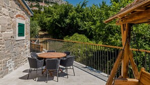 Une terrasse avec une table ronde en bois et des chaises, un mur en pierre et une vue sur une colline parsemée de bâtiments.