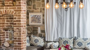 Une pièce chaleureuse avec une cheminée en pierre, des coussins à motifs, une table à manger dressée avec des assiettes et des verres, et un luminaire suspendu.